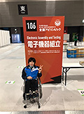 電子機器組立部門の看板の前にいる塩地さんの写真